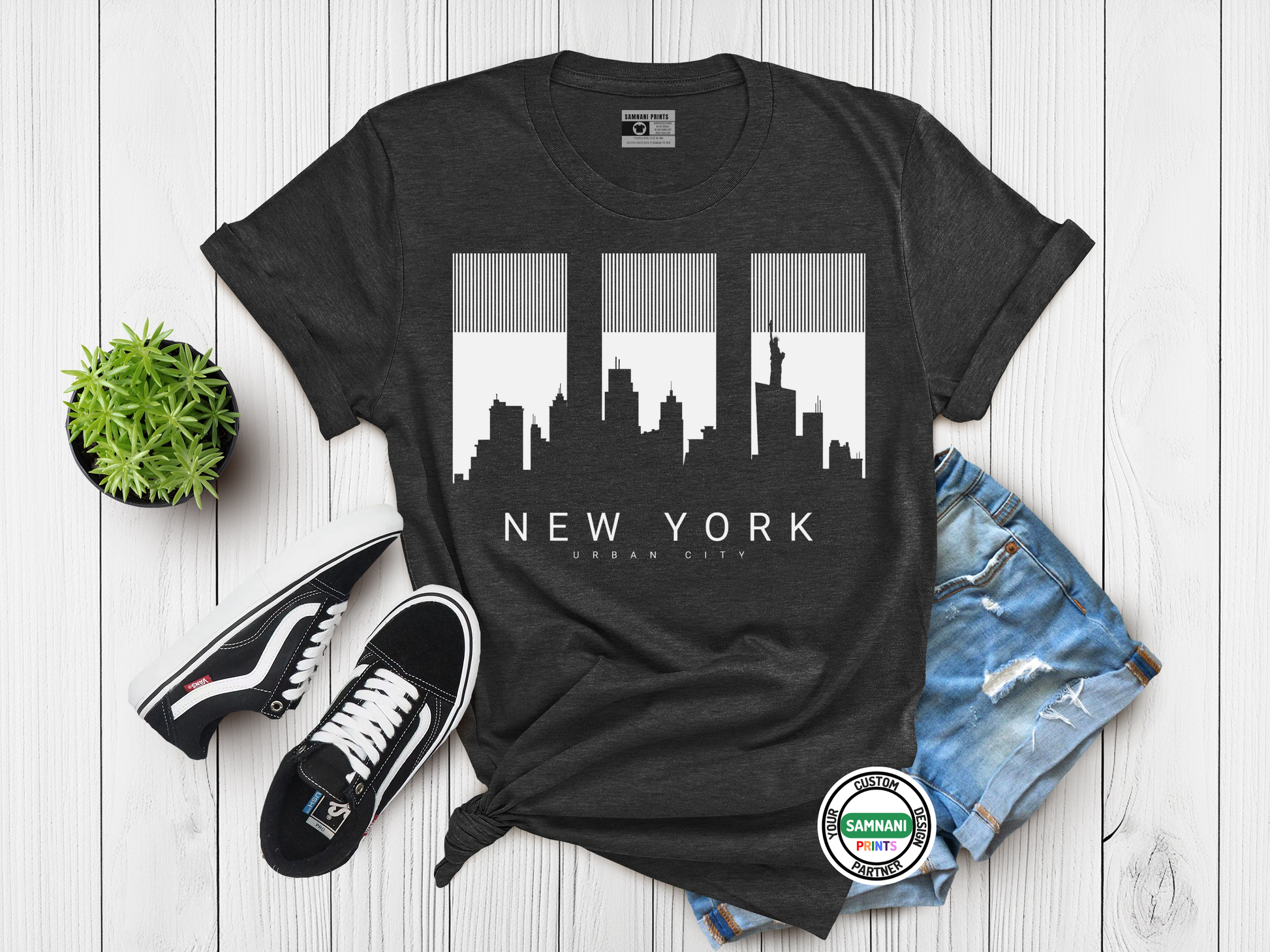 Black t-shirt with New York cityscape design on a wooden surface with shoes and shorts.