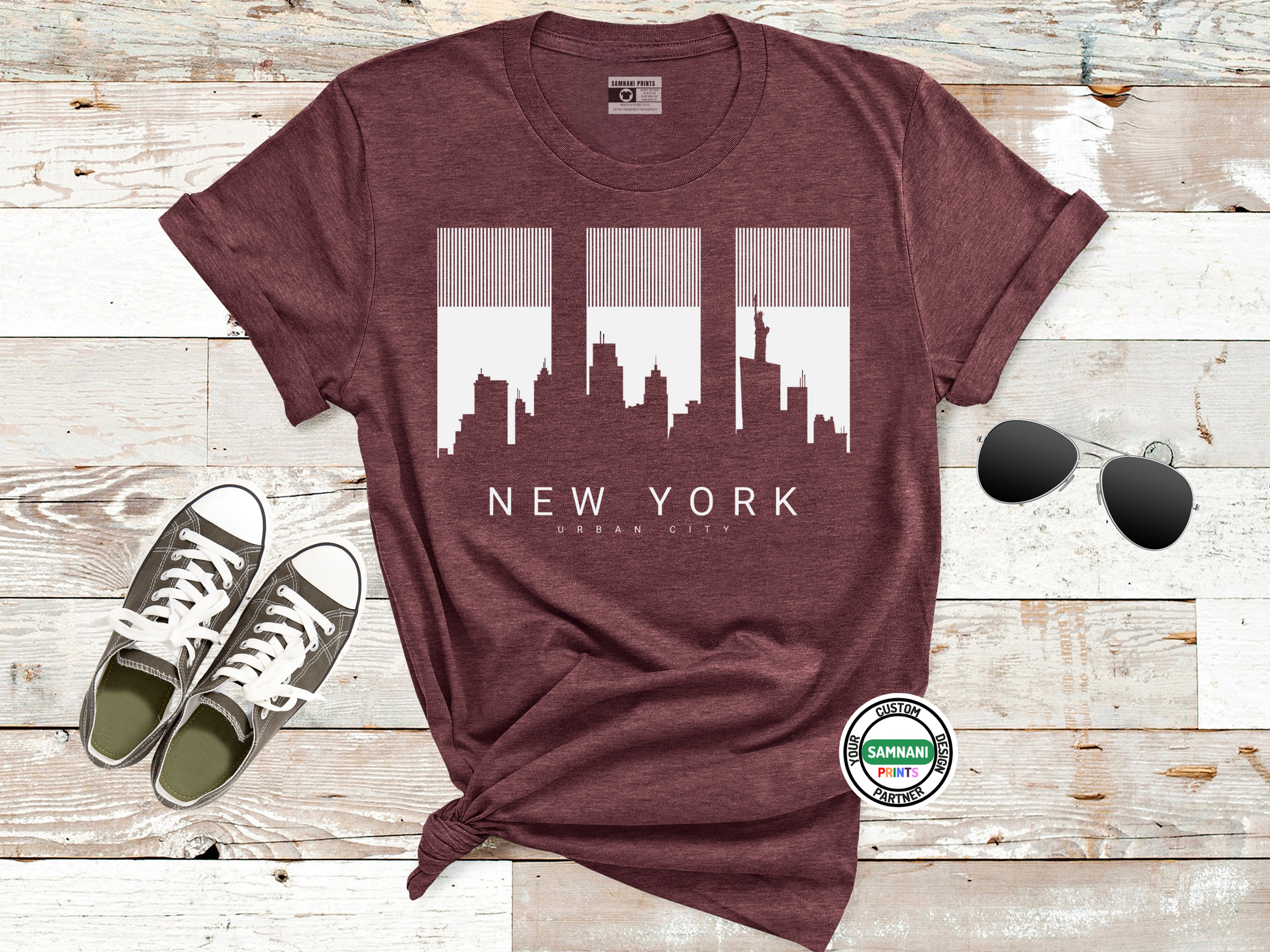 Maroon t-shirt with New York cityscape design on a wooden surface with sneakers and sunglasses.