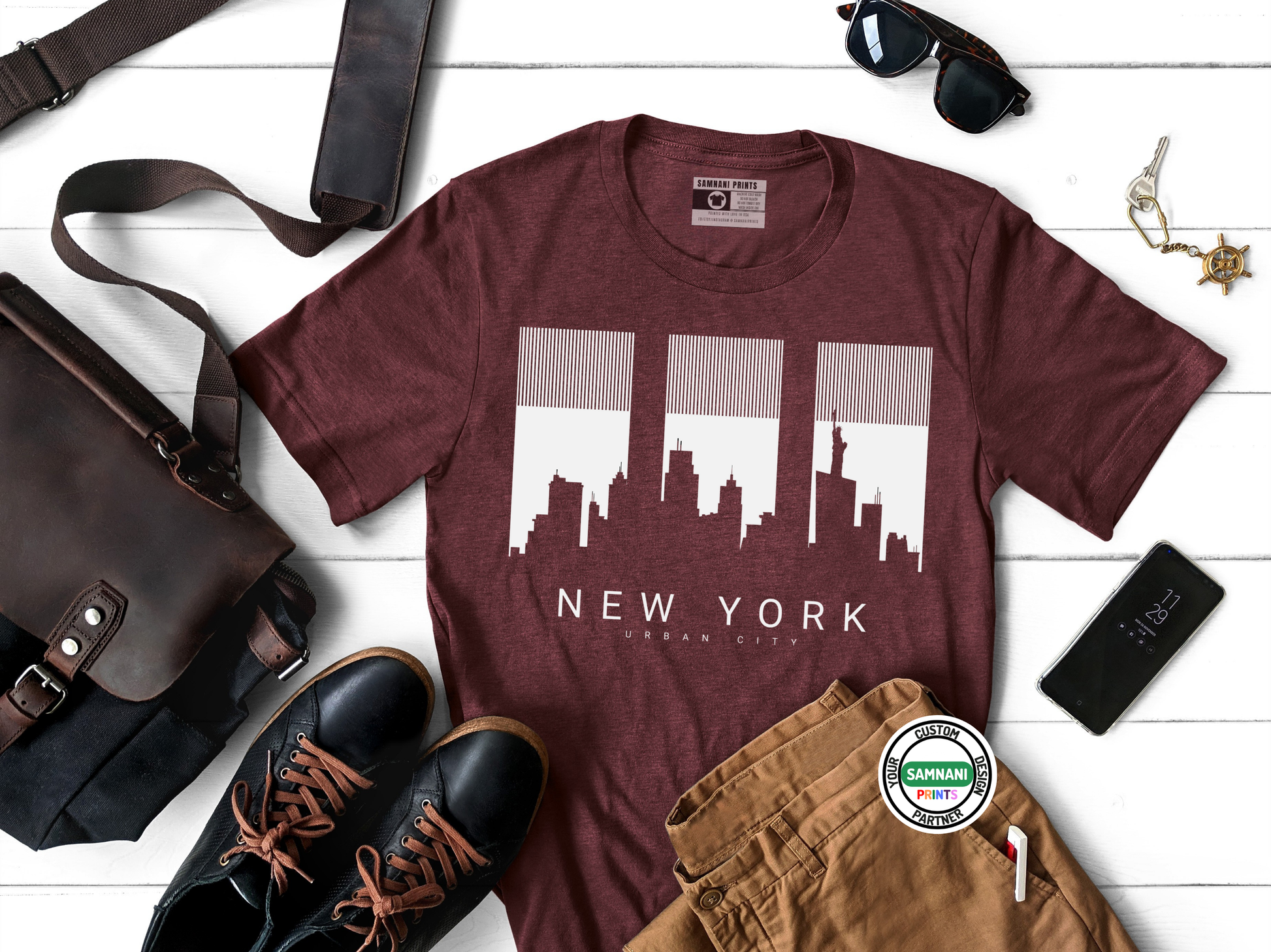 Maroon t-shirt with New York cityscape design, sunglasses, bag, shoes, and pants on a white wooden surface.