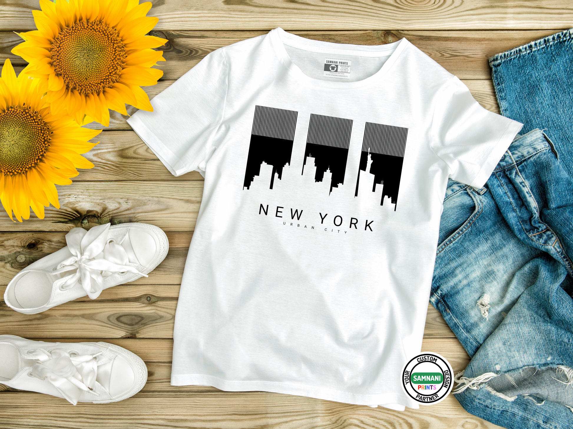 White t-shirt with New York skyline design on a wooden surface with sunflowers, white sneakers, and blue jeans.
