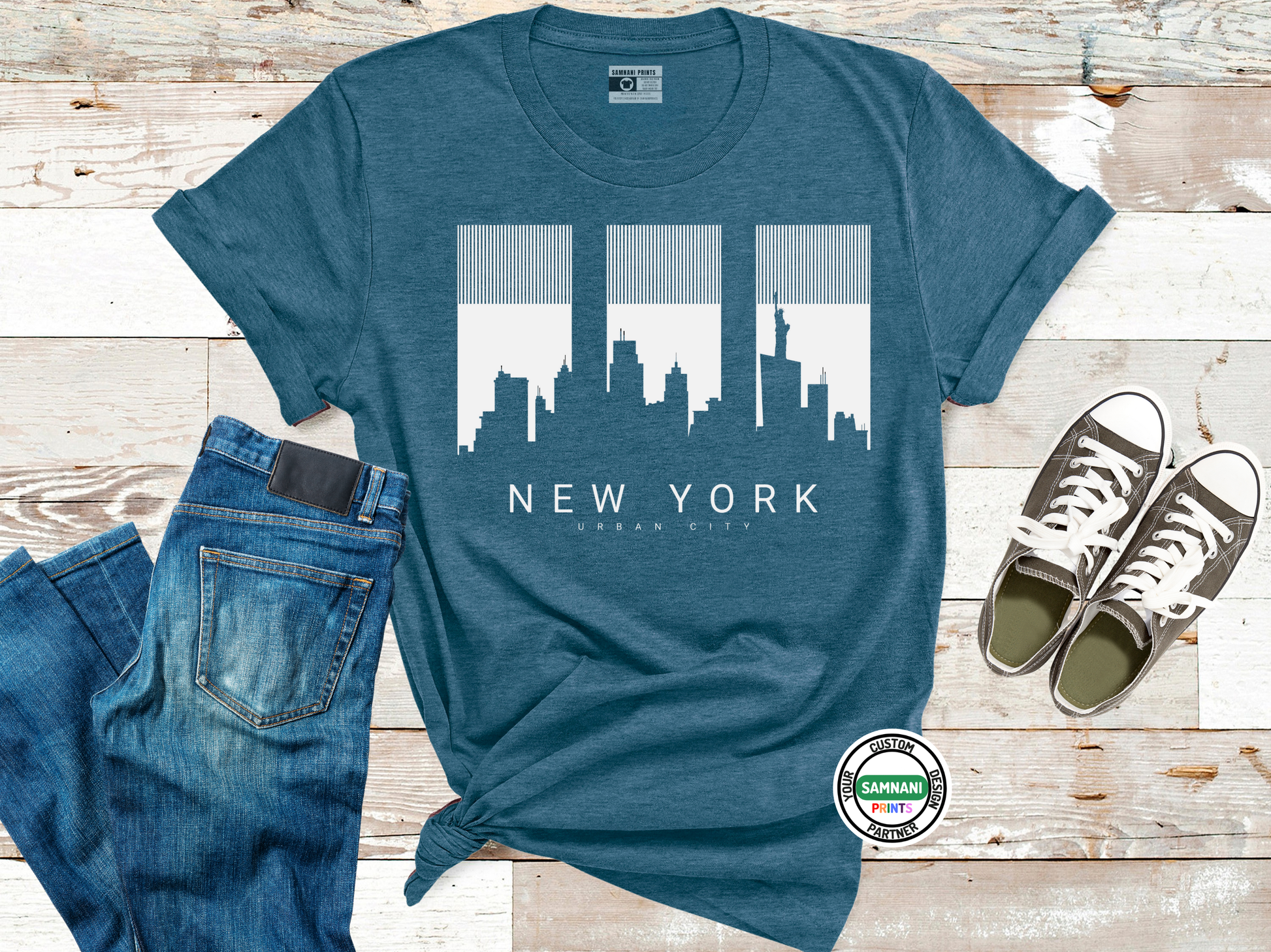 Blue t-shirt with New York cityscape design, jeans, and sneakers on a wooden surface