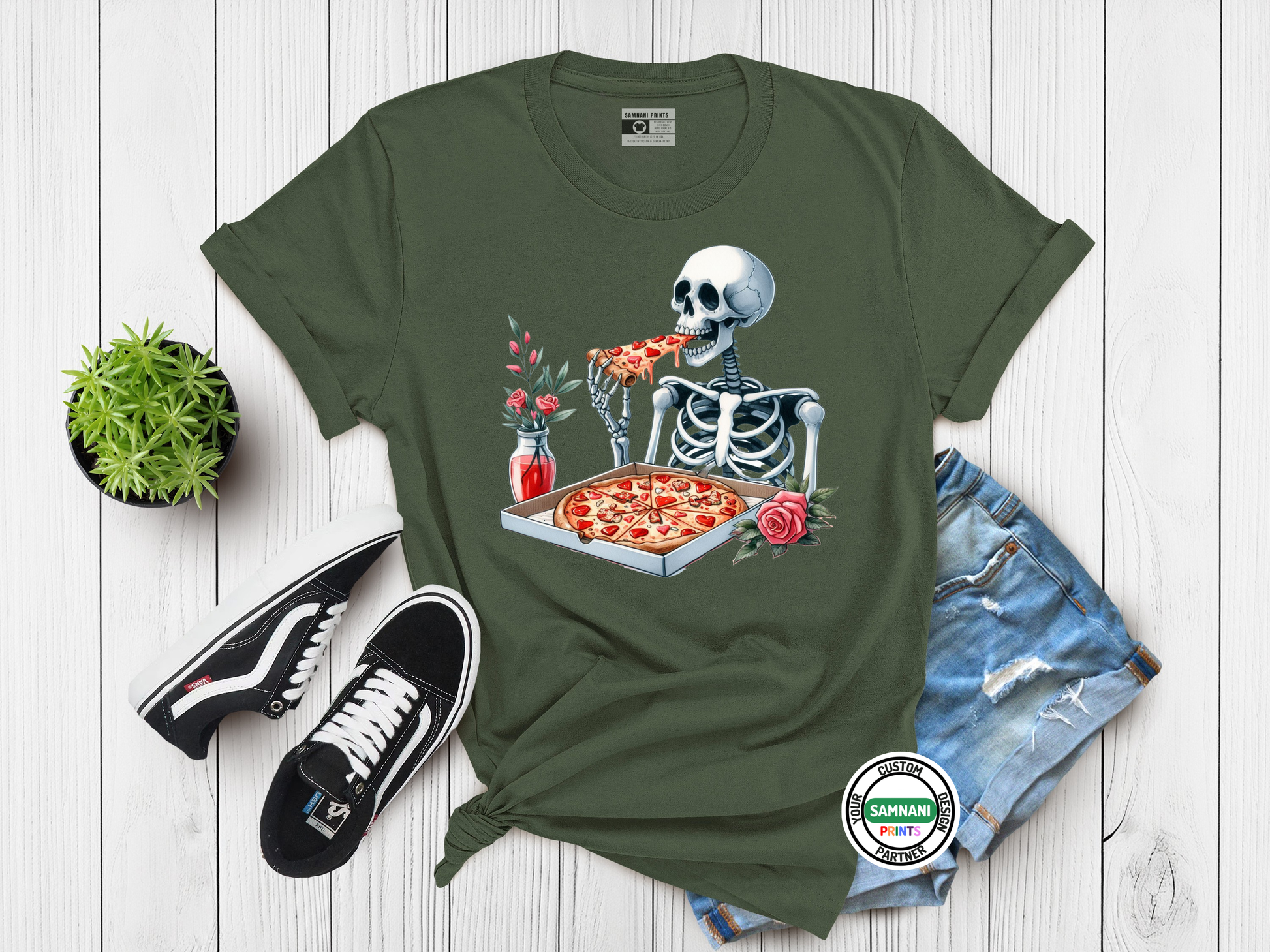 Dark green t-shirt with a detailed graphic of a skeleton eating pizza from a heart-shaped pepperoni box, surrounded by flowers and soda bottle, styled flat with Vans and denim for gothic food lovers.