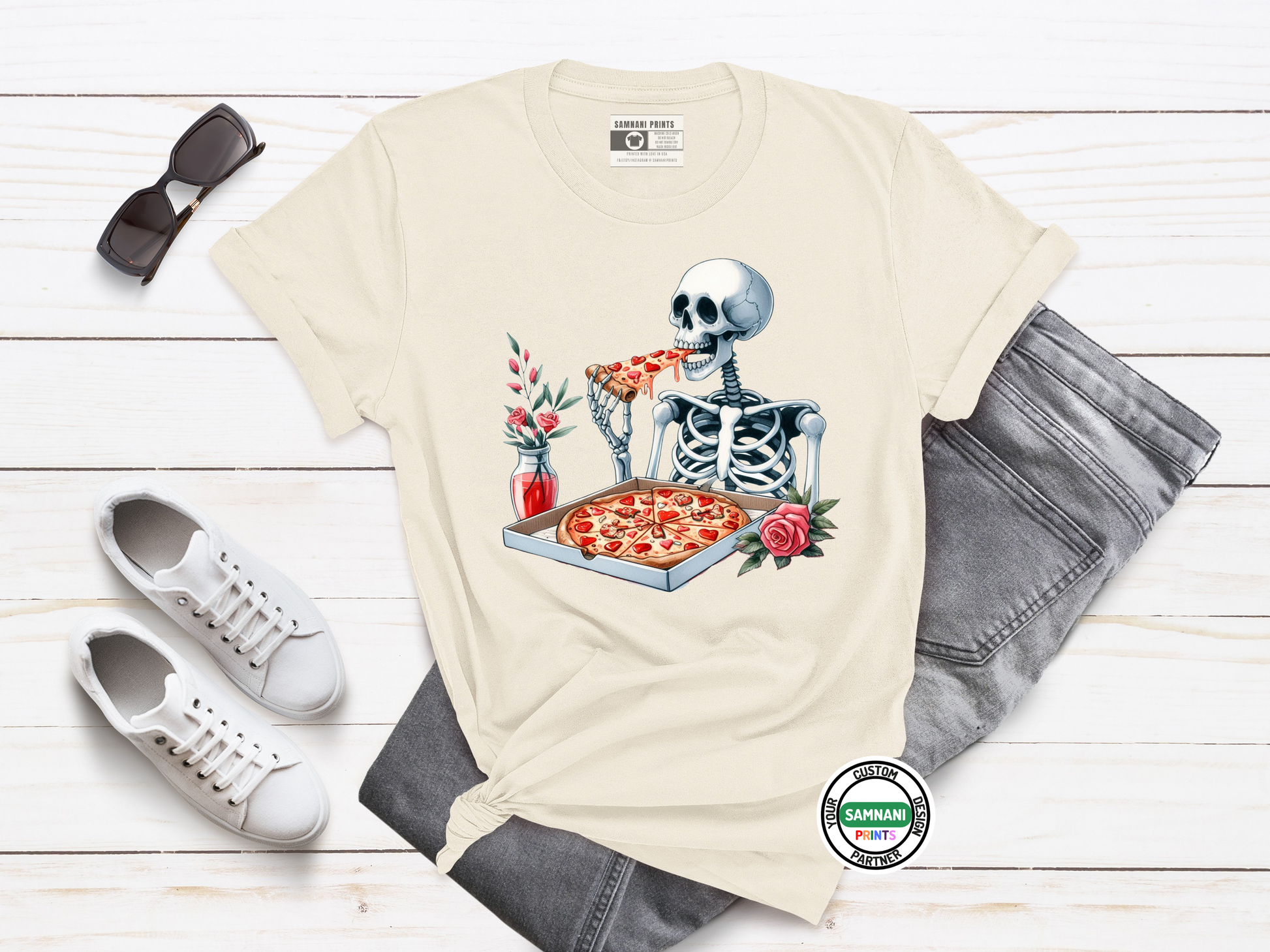 Cream-colored t-shirt with a colorful cartoon skeleton enjoying pizza, surrounded by romantic elements like roses and soda, styled with gray denim, ideal for quirky Halloween or foodie fashion.