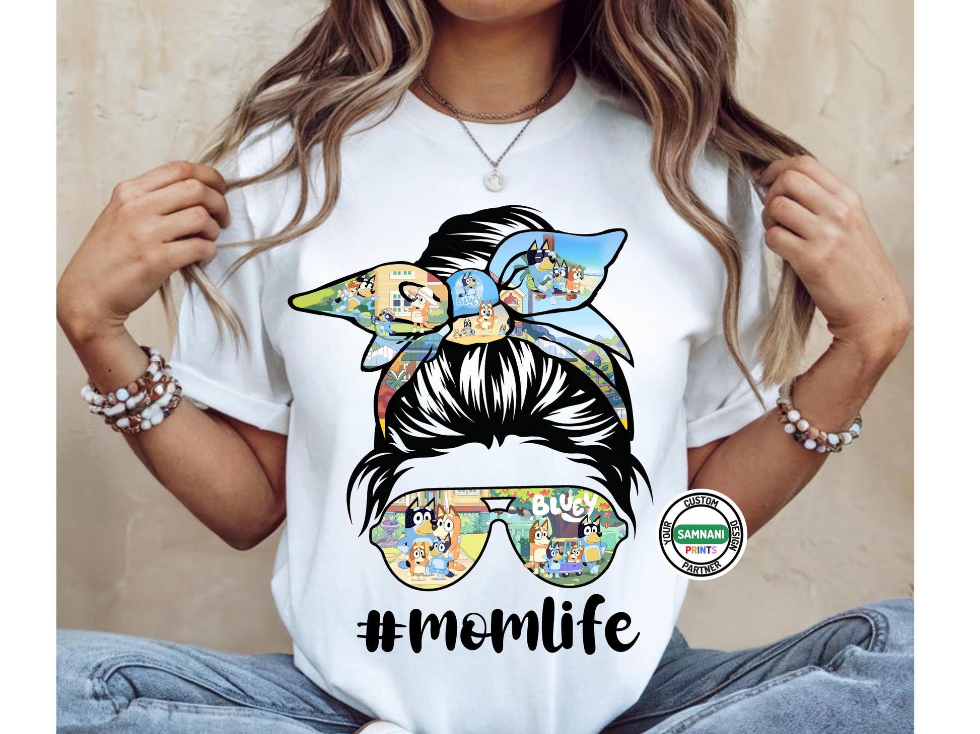 A woman wearing a stylish white "Mom Life" t-shirt with a messy bun graphic featuring a Bluey-themed bow and sunglasses. The shirt includes the hashtag #MomLife, making it a fun and trendy choice for moms who love cartoons and casual fashion. She is accessorized with bracelets and a necklace, completing the relaxed and modern look.