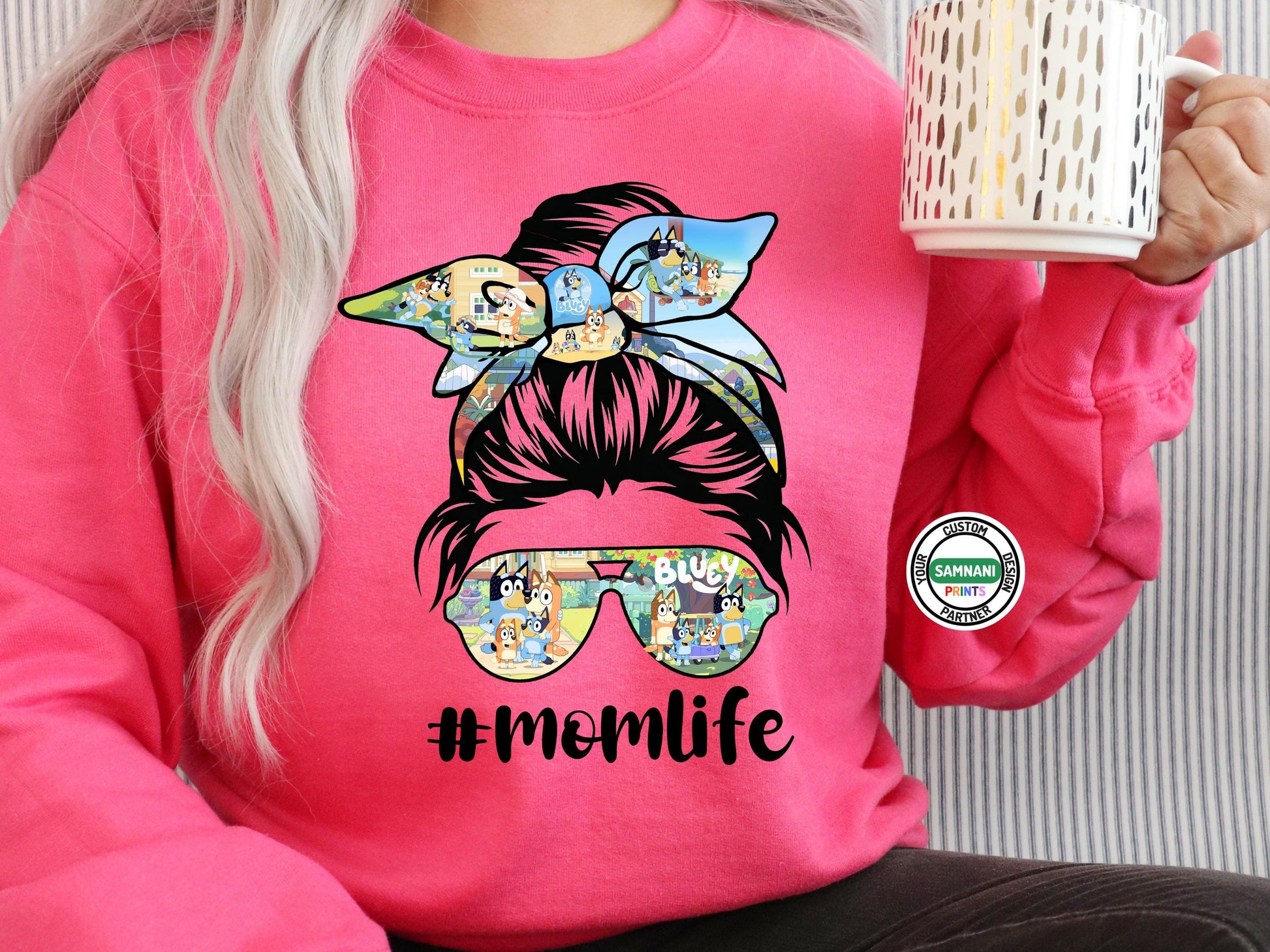 A woman dressed in a comfy and trendy "Mom Life" t-shirt, showcasing a messy bun graphic with a Bluey-themed bow and sunglasses. The fun #MomLife design makes it a great gift for cartoon-loving moms or those embracing motherhood in style. She pairs the look with bracelets and a necklace, adding a fashionable touch.