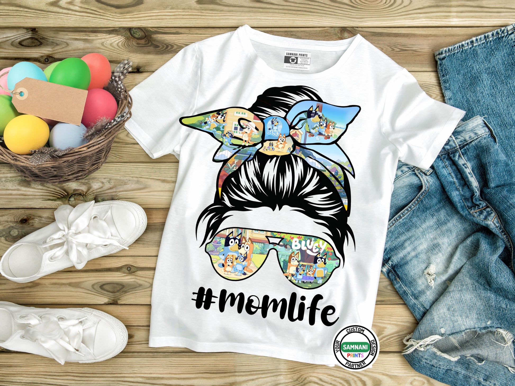 A flat lay t shirt a trendy white "Mom Life" t-shirt featuring a messy bun with a Bluey-themed bow and sunglasses, along with the hashtag #MomLife in black text. The stylish and fun design makes it a perfect casual outfit for moms who love cartoons and playful fashion. The woman is accessorized with bracelets and a necklace, adding to the trendy aesthetic.