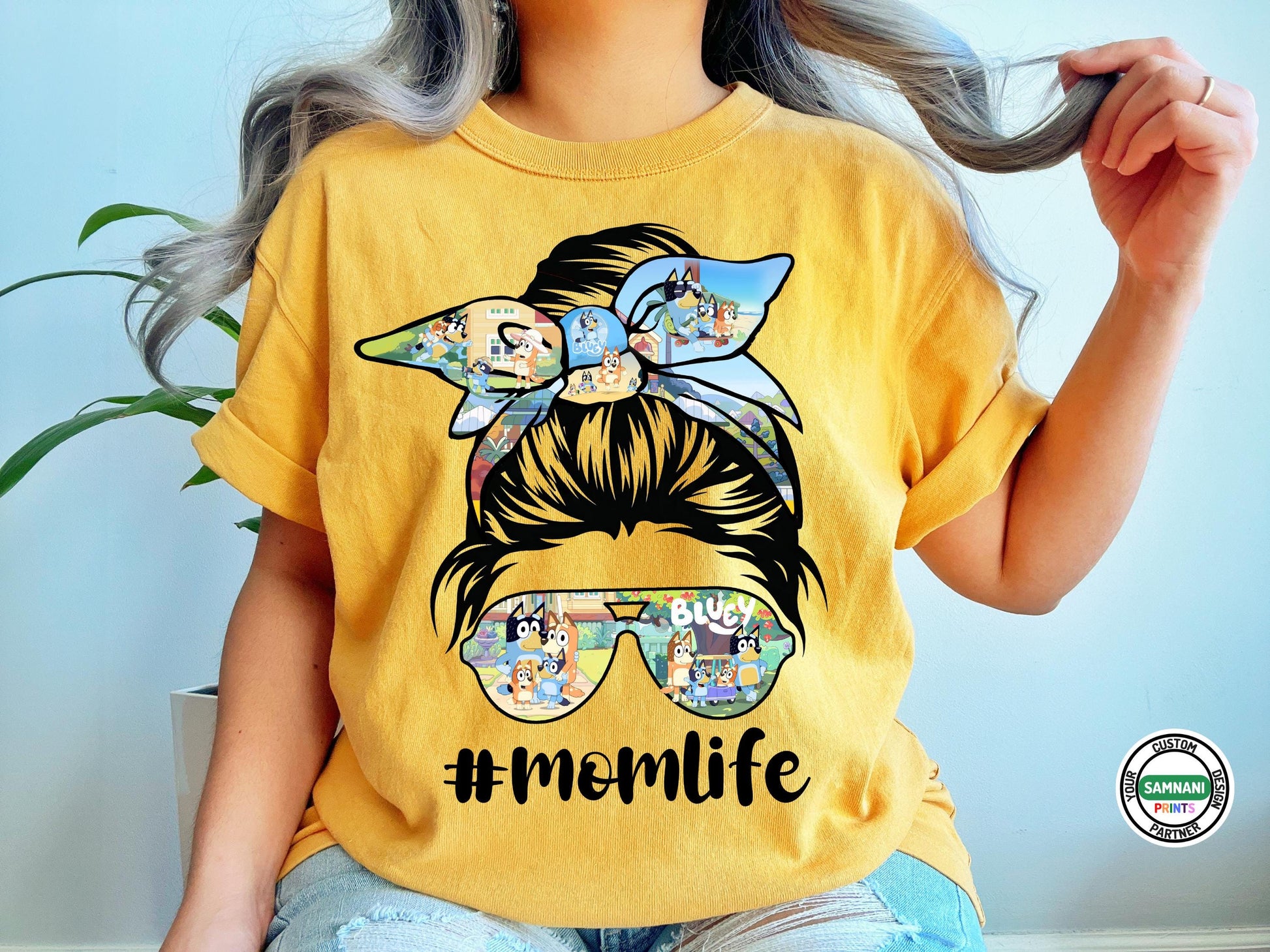 A fashionable mom wearing a white "Mom Life" graphic tee featuring a messy bun design with a Bluey-inspired bow and sunglasses. The playful #MomLife print adds a trendy touch, perfect for casual wear, playdates, or everyday outings. She is accessorized with jewelry, enhancing the stylish and effortless mom aesthetic.