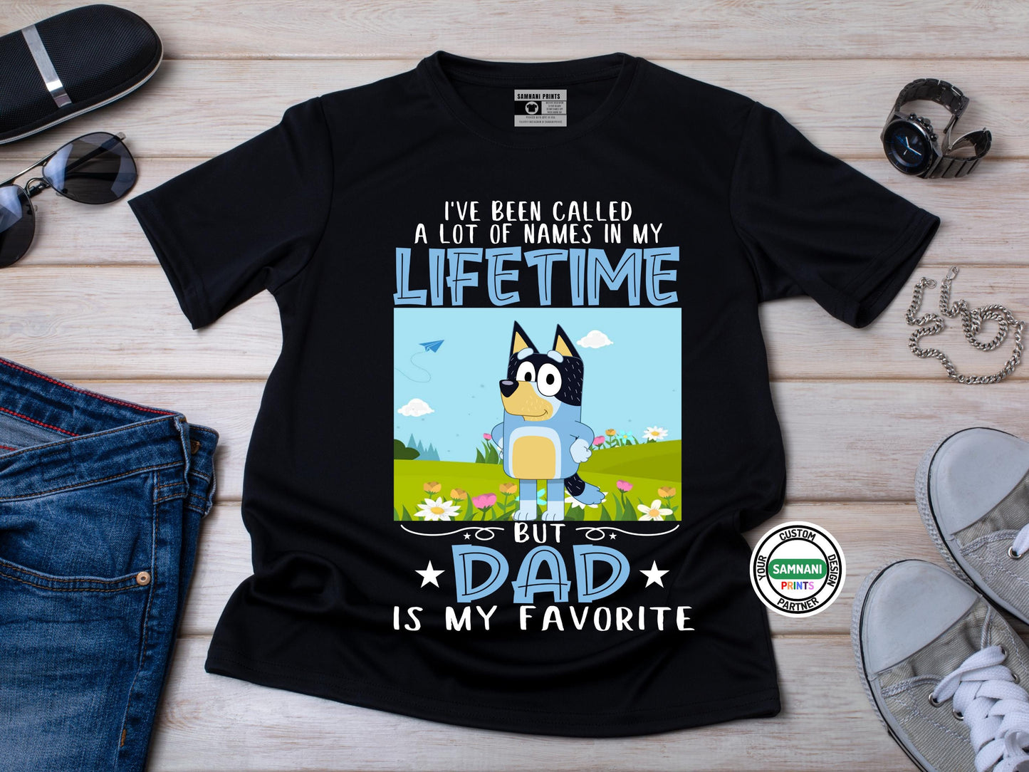 A flat lay Black t-shirt with a cartoon dad character in a field of flowers and text that reads “I’ve Been Called a Lot of Names in My Lifetime, But Dad Is My Favorite.” Bluey Dad Shirt – I've Been Called a Lot of Names in My Lifetime But Dad Is My Favorite – Funny Father's Day Gift. Father's Day gift, gift for dad, gift for paw paw, gift for father. Bluey Bandit T-shirt and sweatshirt.