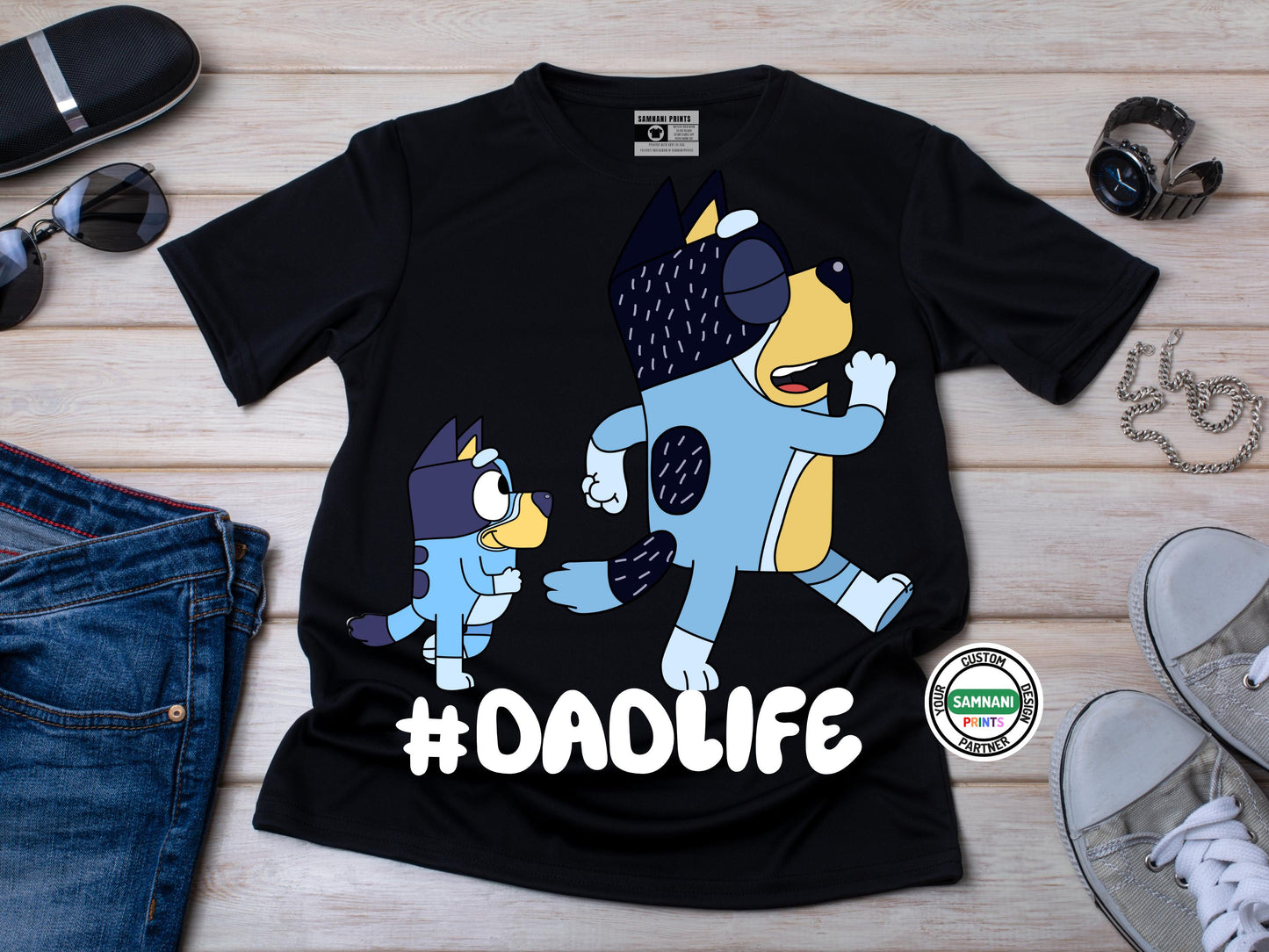 A flat lay black t-shirt with cartoon dad and a kid graphic above the hashtag "#DADLIFE," styled with black shoes, bag, sunglasses, and brown pant. Father's Day gift, gift for dad under $20, gift for dad, Bluey and Bandit T-shirt and sweatshirt, Father and daughter tee, gift for father.