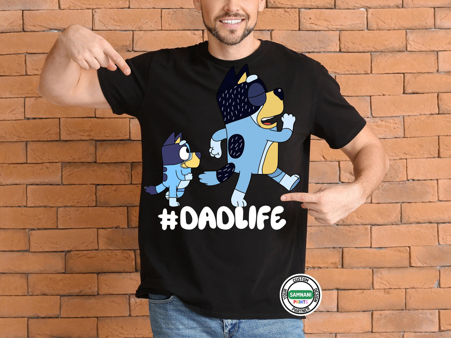 man wearing a black t-shirt with cartoon dad and a kid graphic above the hashtag "#DADLIFE," smiling and pointing at the design, standing against s brick wall pointing at shirt. Father's Day gift, gift for dad under $20, gift for dad, Bluey and Bandit T-shirt and sweatshirt, Father and daughter tee, gift for father.