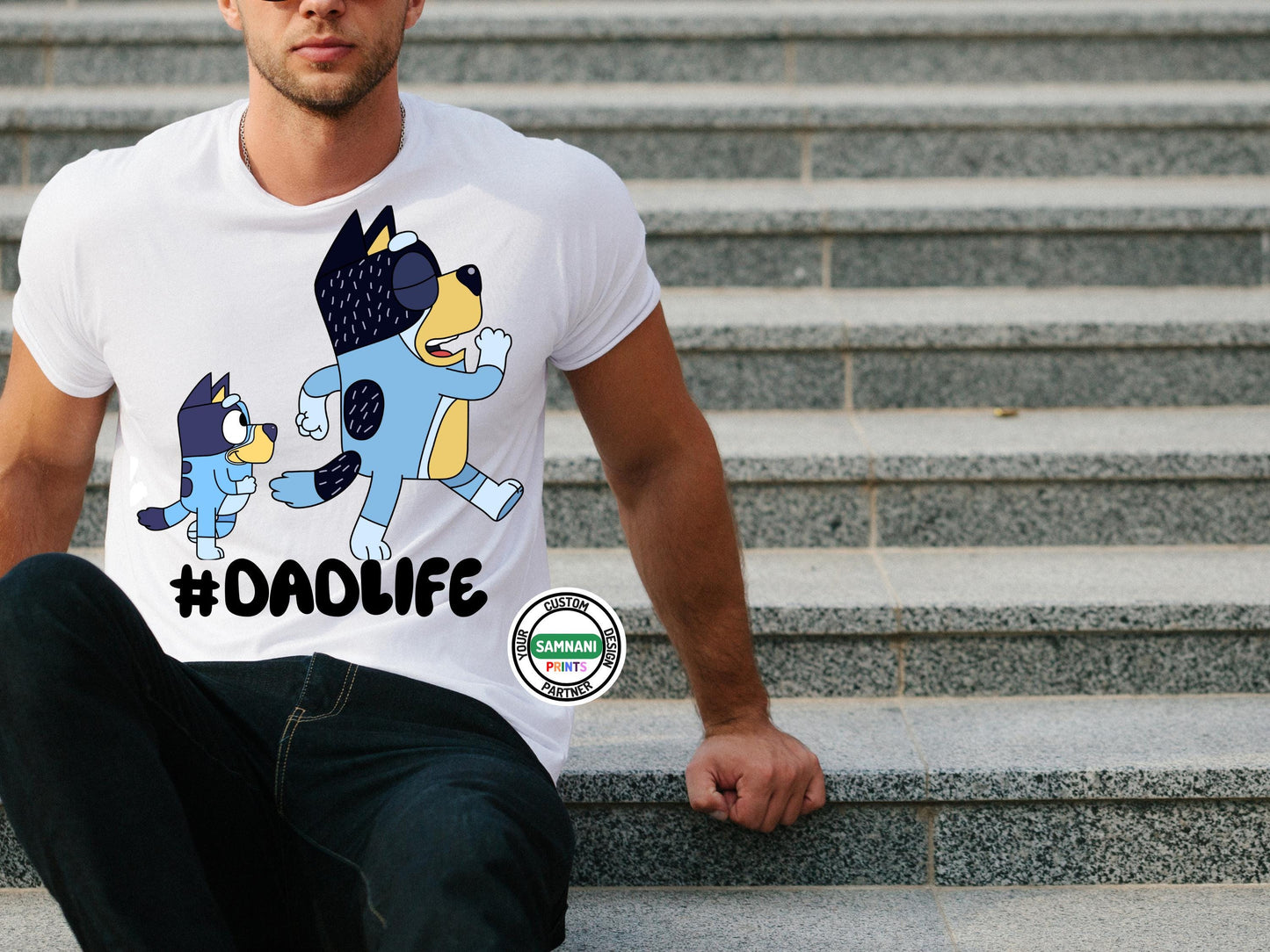 Man wearing a white t-shirt with a blue and black cartoon dog design. The shirt has the text '#DADLIFE' printed on the front sitting on stairs.