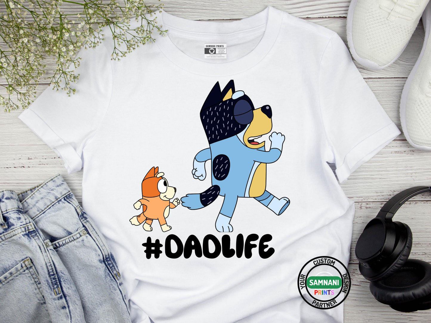 A flat lay white t-shirt with cartoon dad and a kid graphic above the hashtag "#DADLIFE," smiling and pointing at the design, standing against a brick wall background. Bingo and Bandit Bluey inspired t-shirt, Bluey dad gift, Father's Day gift from daughter to dad. Father's Day gift for Bluey fans. Father's Day gift under $20.