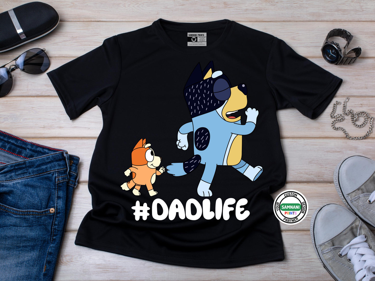 A flat lay black t-shirt with cartoon dad and a kid graphic above the hashtag "#DADLIFE," smiling and pointing at the design, standing against a brick wall background. Bingo and Bandit Bluey inspired t-shirt, Bluey dad gift, Father's Day gift from daughter to dad. Father's Day gift for Bluey fans. Father's Day gift under $20.
