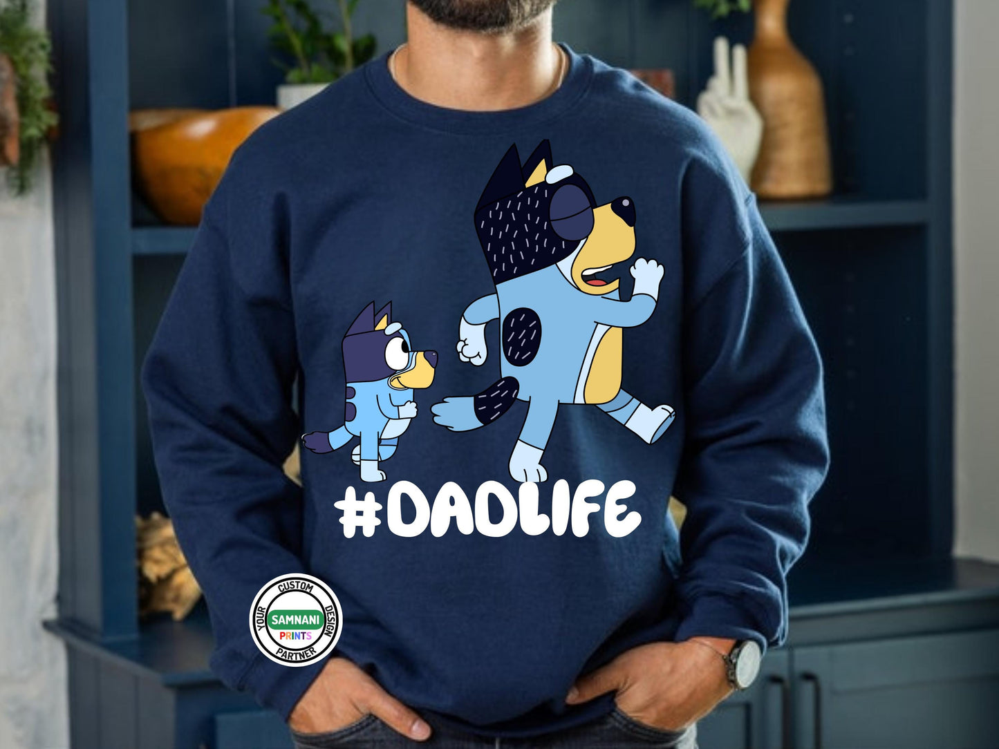 Man wearing a navy blue sweatshirt with a graphic of two cartoon dogs. The dogs are blue and yellow with black markings. The larger dog is wearing a blue and yellow outfit and the smaller dog is wearing a blue and yellow outfit. The text '#DADLIFE' is printed in white below the dogs.