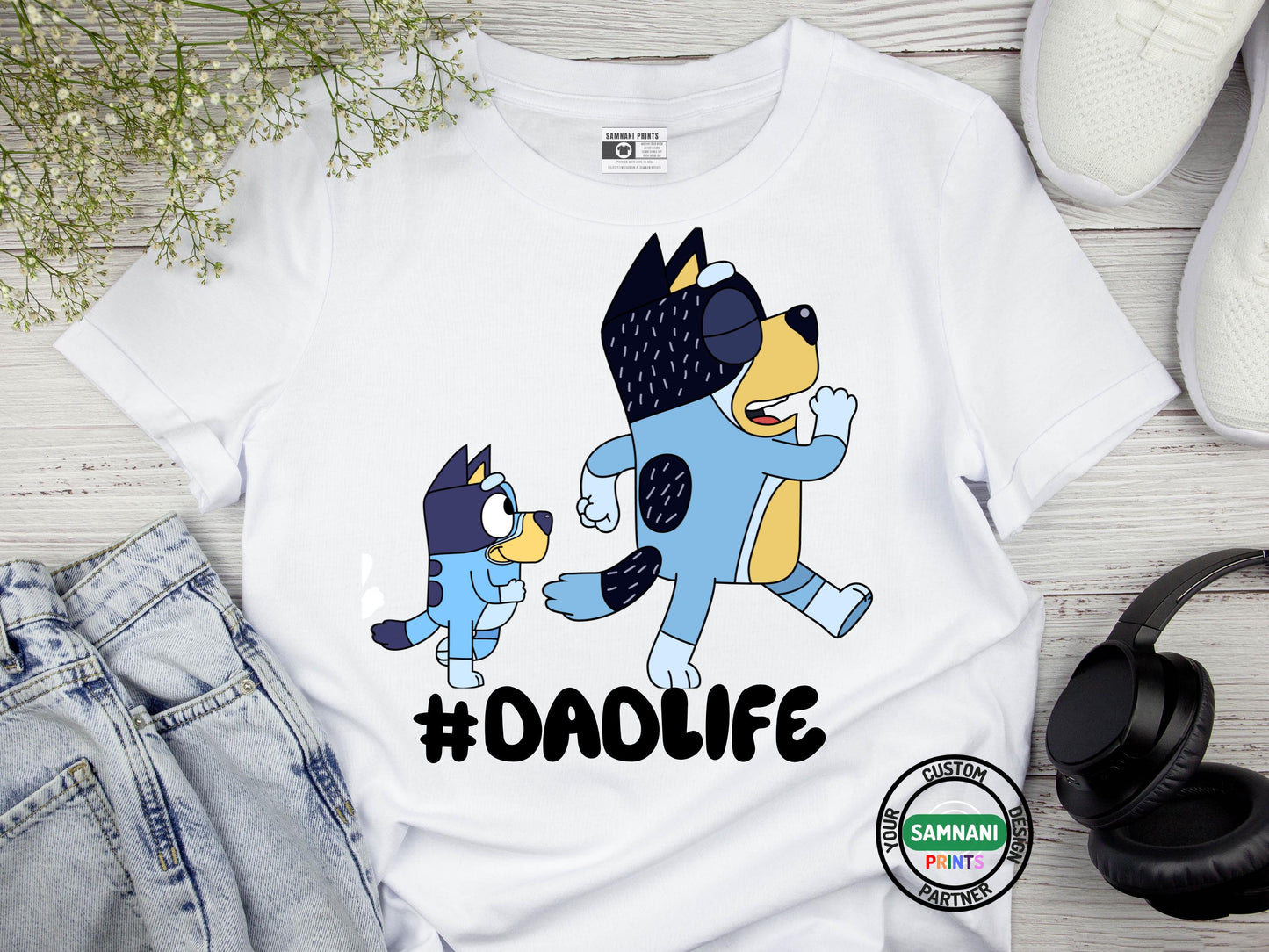 A flat lay white t-shirt with cartoon dad and a kid graphic above the hashtag "#DADLIFE," smiling and pointing at the design, styled with denim jeans white shoes, and headphones. Father's Day gift, gift for dad under $20, gift for dad, Bluey and Bandit T-shirt and sweatshirt, Father and daughter tee, gift for father.