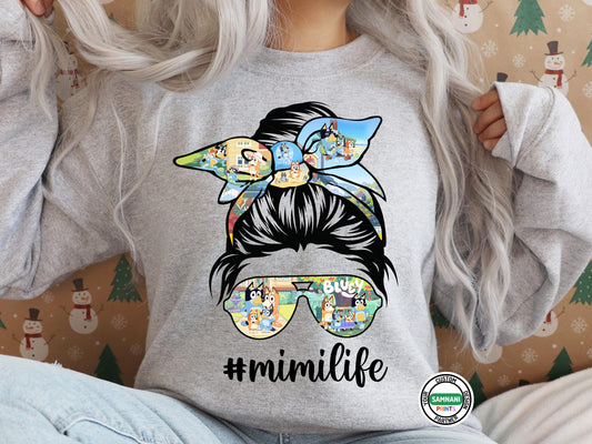 Women wearing a grey sweatshirt featuring a cartoon-themed messy bun design with the hashtag #mimilife. With a Christmas wallpaper background #mimilife Bluey