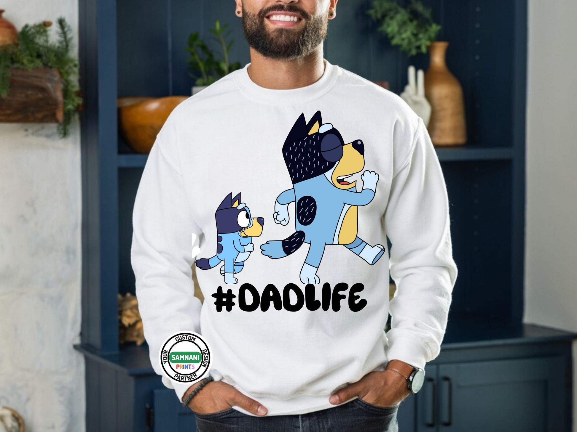 Man wearing a white sweatshirt with cartoon dad and a kid graphic above the hashtag &quot;#DADLIFE,&quot; smiling and pointing at the design, standing against s brick wall pointing at shirt. Father&#39;s Day gift, gift for dad under $20, gift for dad, Bluey and Bandit T-shirt and sweatshirt, Father and daughter tee, gift for father.